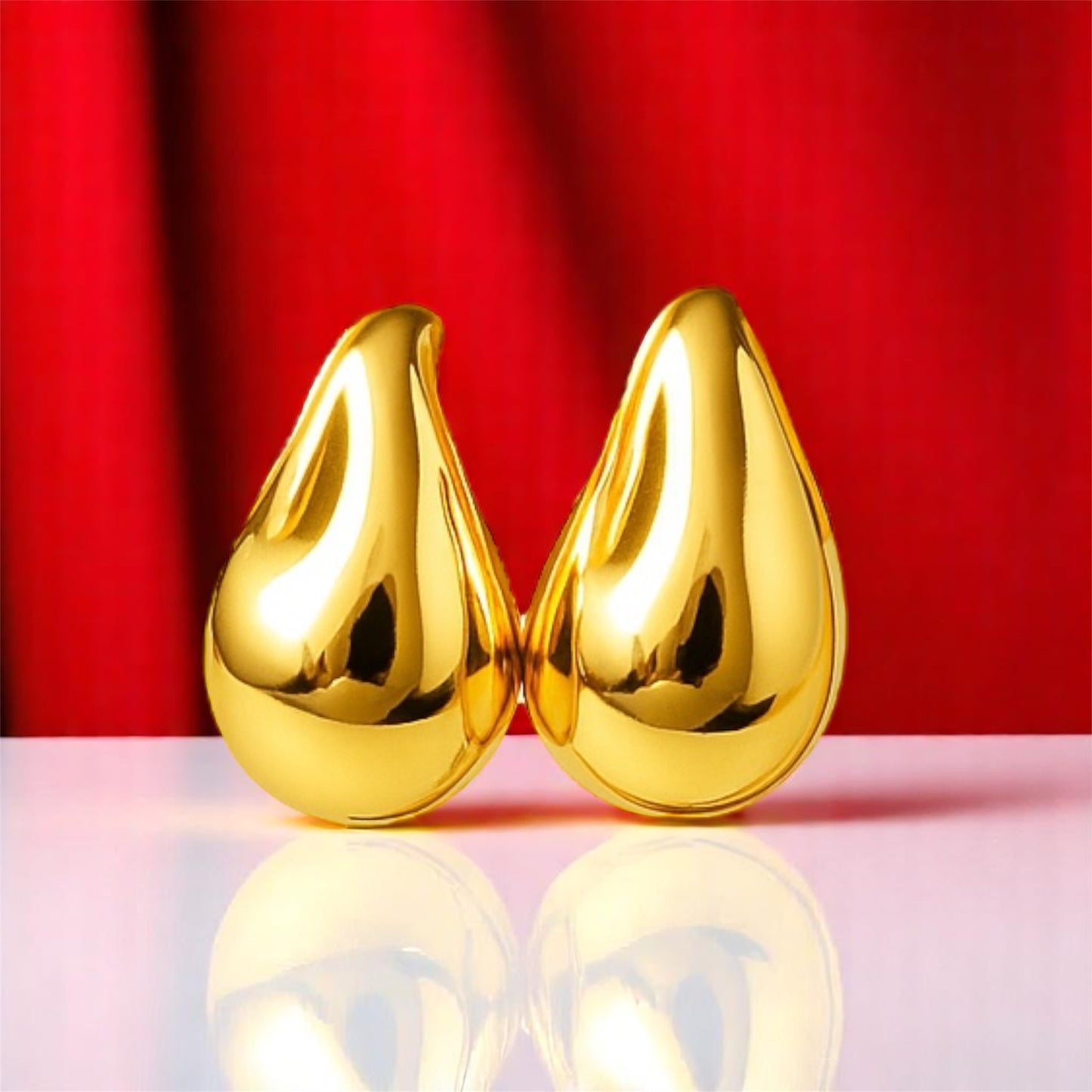 Two gold teardrop-shaped objects on a reflective surface with a red curtain background