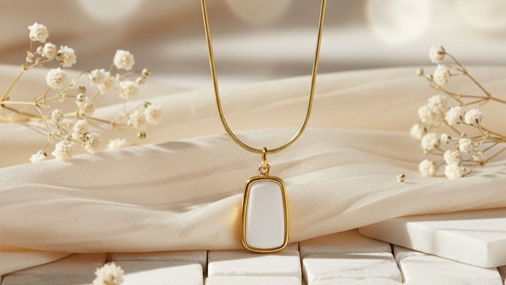 Gold necklace with a white pendant on a textured beige surface with dried flowers.