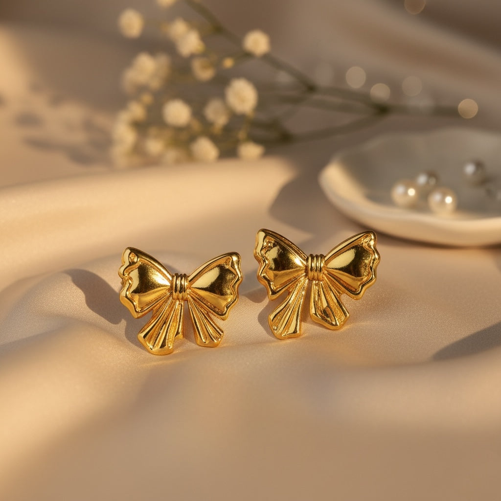 Gold bow-shaped earrings on a beige surface with blurred flowers in the background